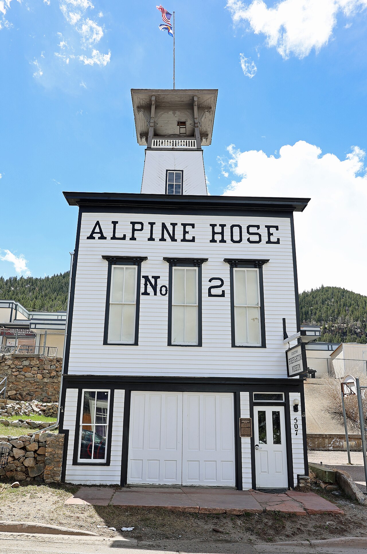 A narrow white firehouse lettered Alpine Hose No. 2 between three tall windows, double doors below and a tall square bell tower behind.