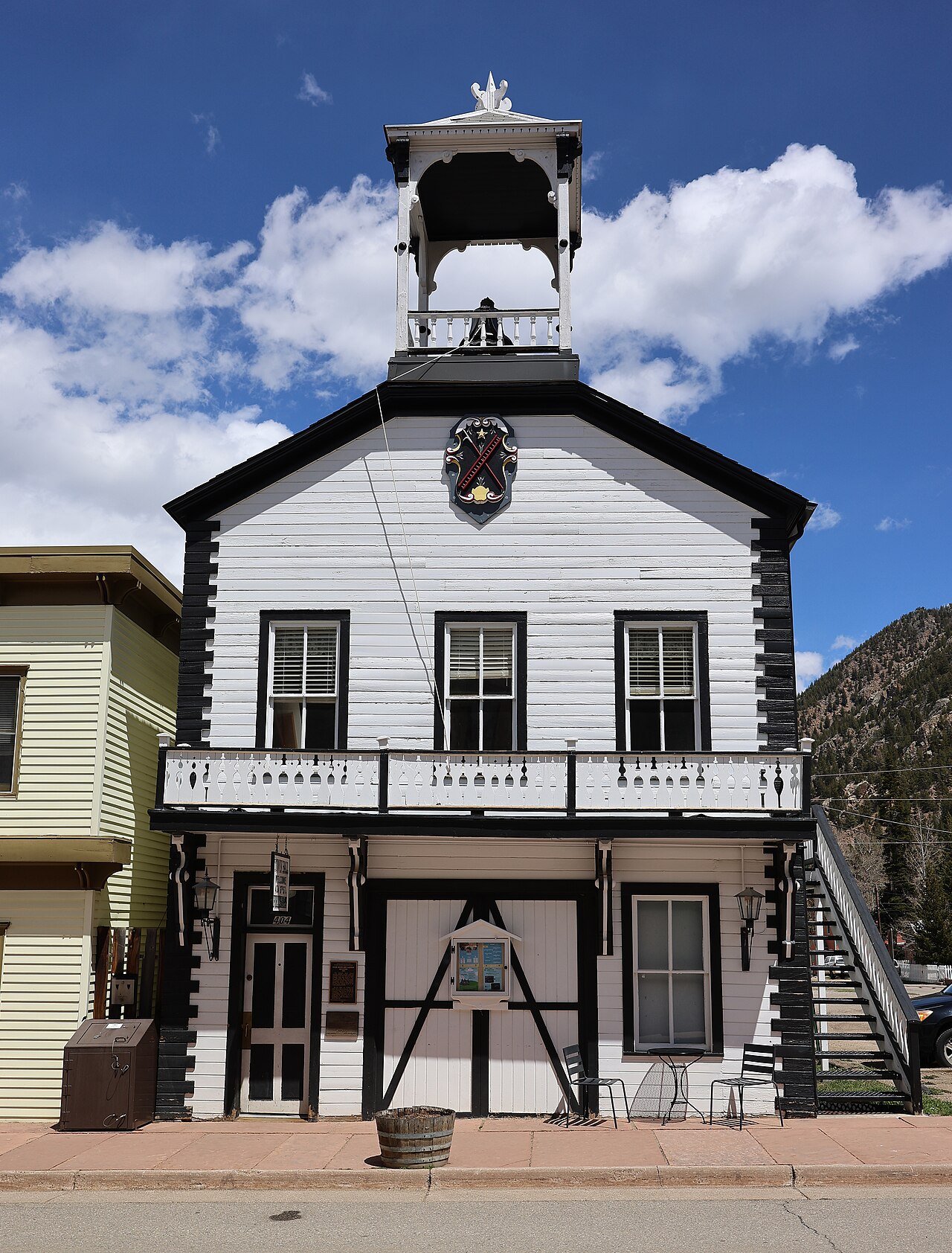 A white clapboard two-story building with a bell tower on the roof, a fire company shield in the gable, a long balcony and wide equipment doors.