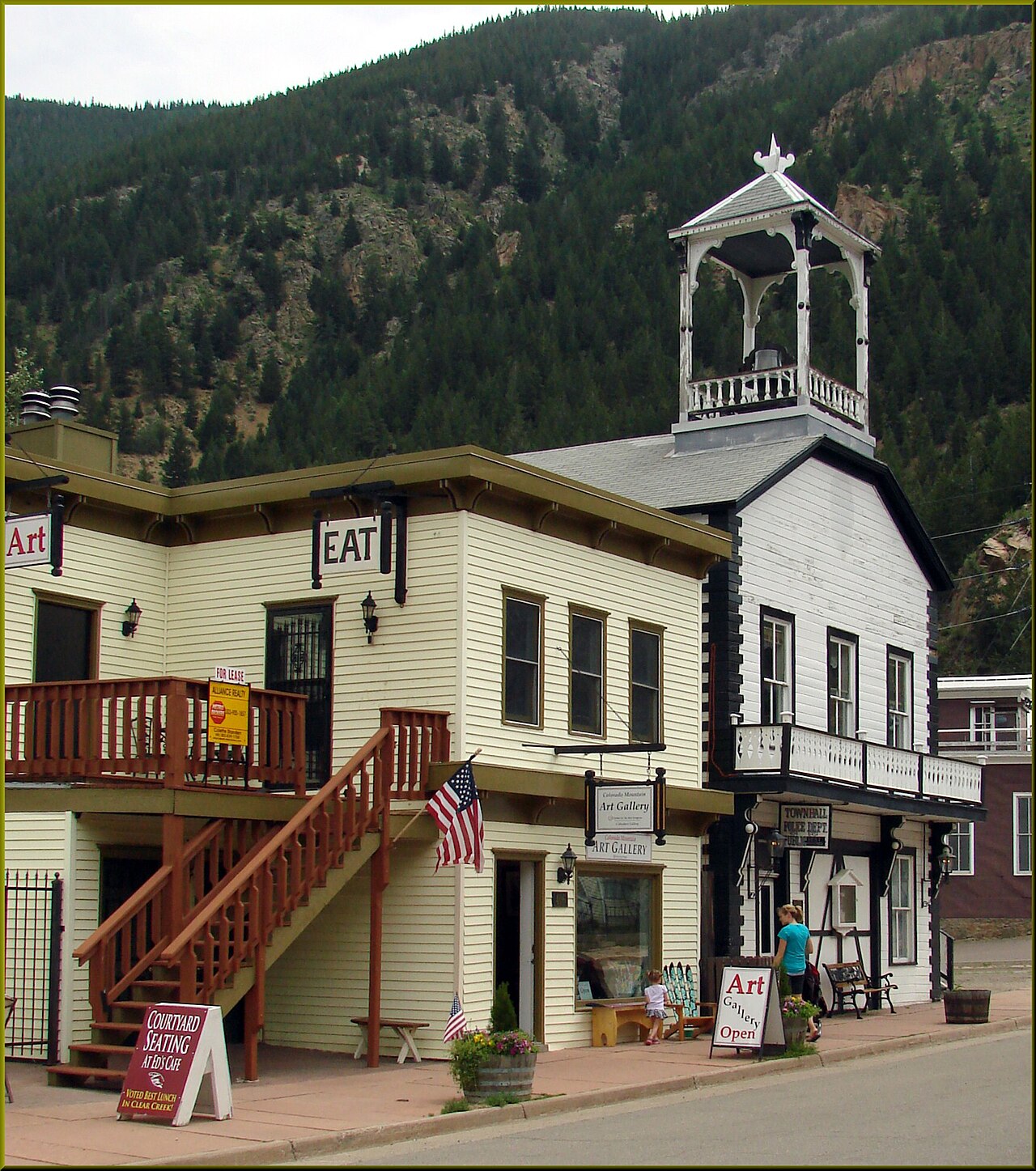 A white wooden building with a small open bell tower, seen from the street beside a neighboring storefront.