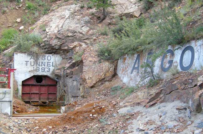 A small arched opening closed by a rust-red door in a rock face, the words Argo Tunnel 1893 painted above.