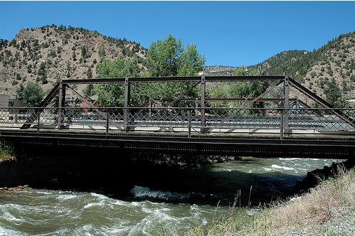 A low dark steel truss bridge over a fast mountain creek, wooded hills behind.