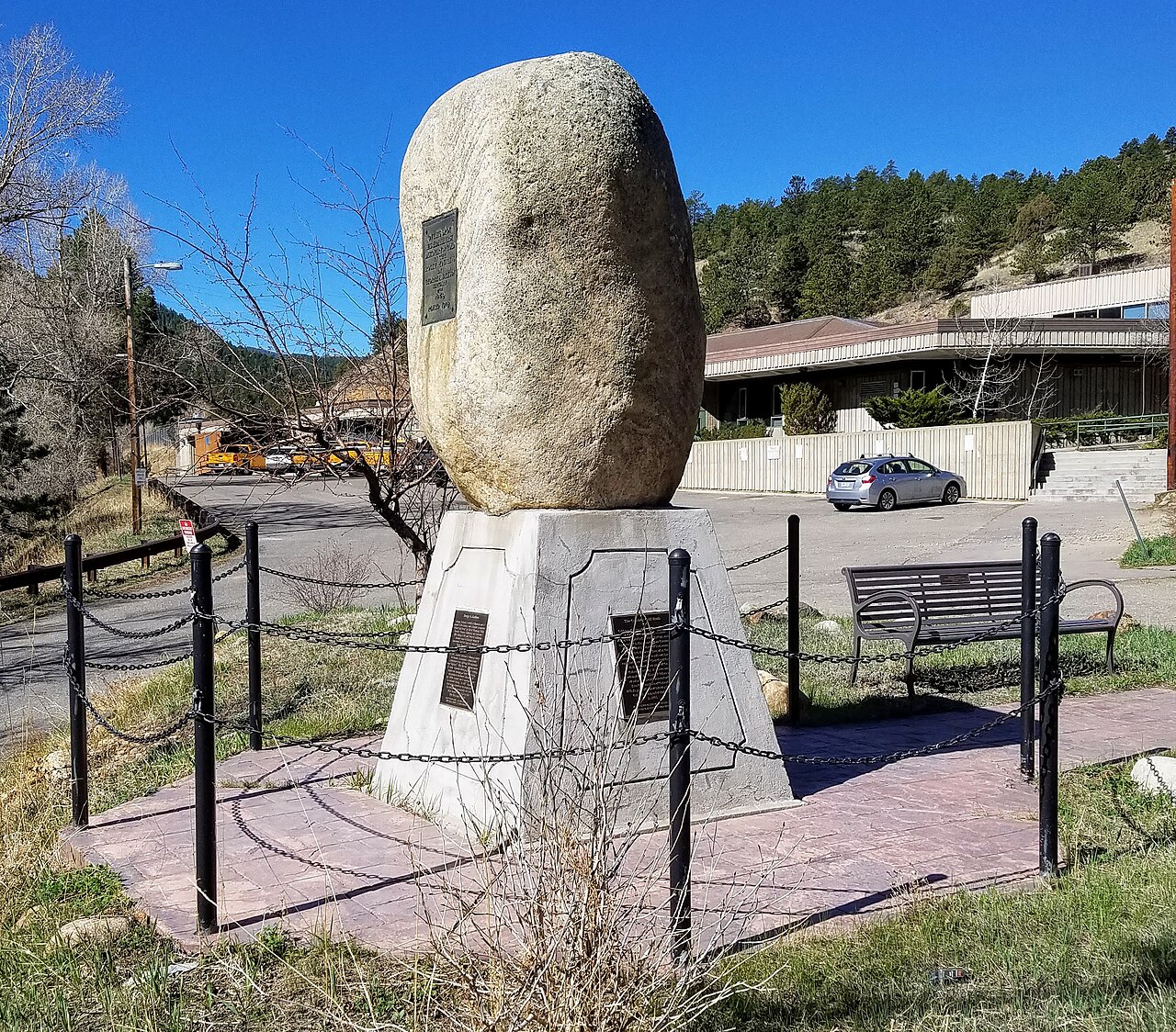 A large rounded boulder on a white pedestal with plaques, inside a chain fence.