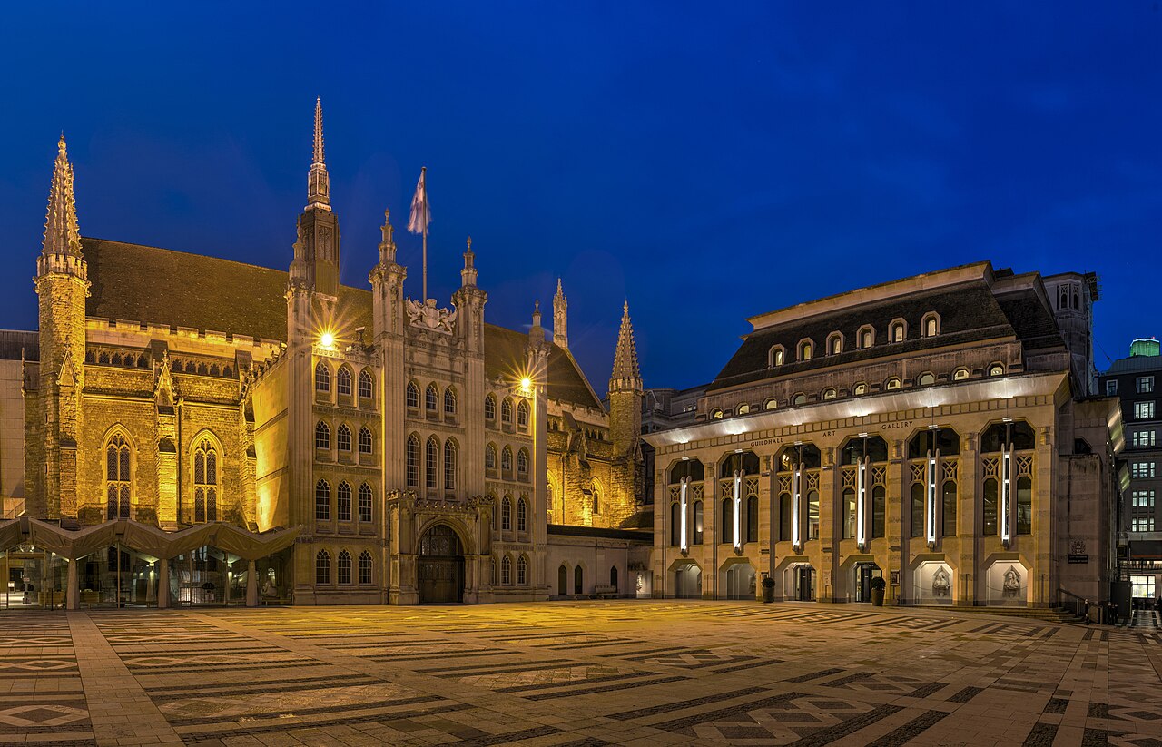 A floodlit medieval hall with a pinnacled porch across a wide paved yard at blue hour.