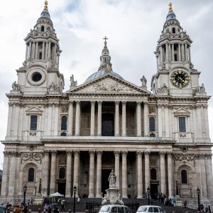 A domed baroque cathedral with two towers and a columned portico, a statue on a pedestal in front, taxis on the road.