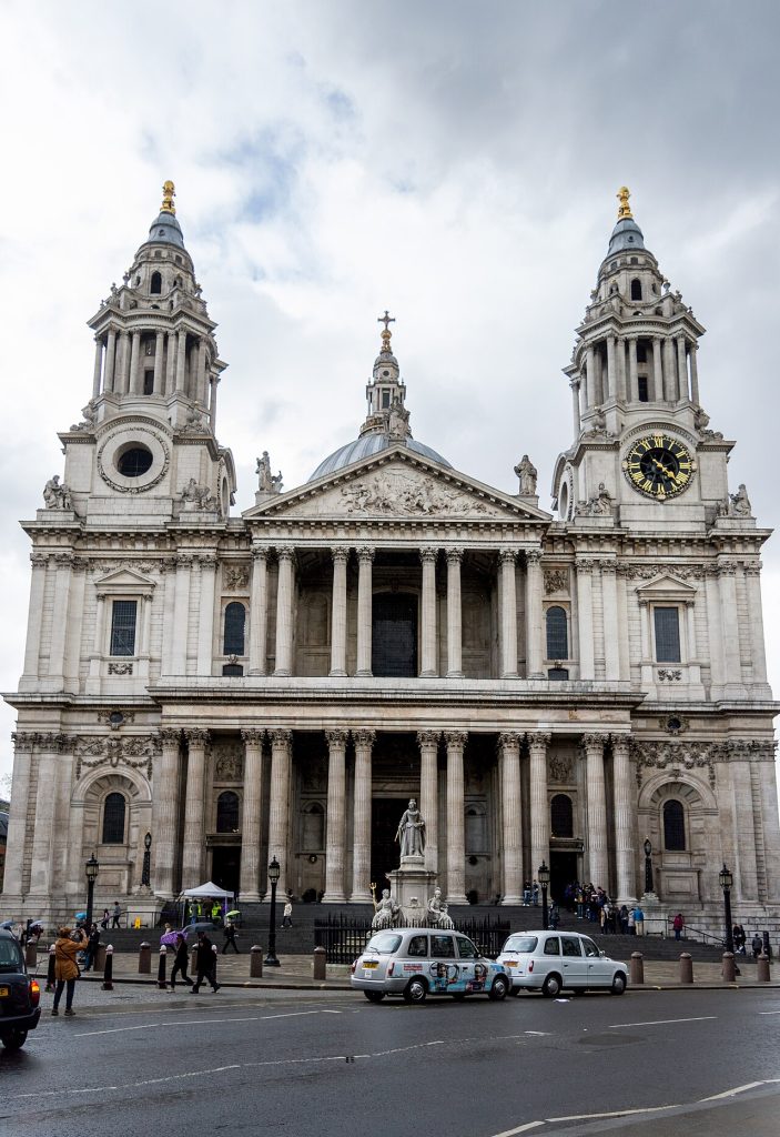 A domed baroque cathedral with two towers and a columned portico, a statue on a pedestal in front, taxis on the road.