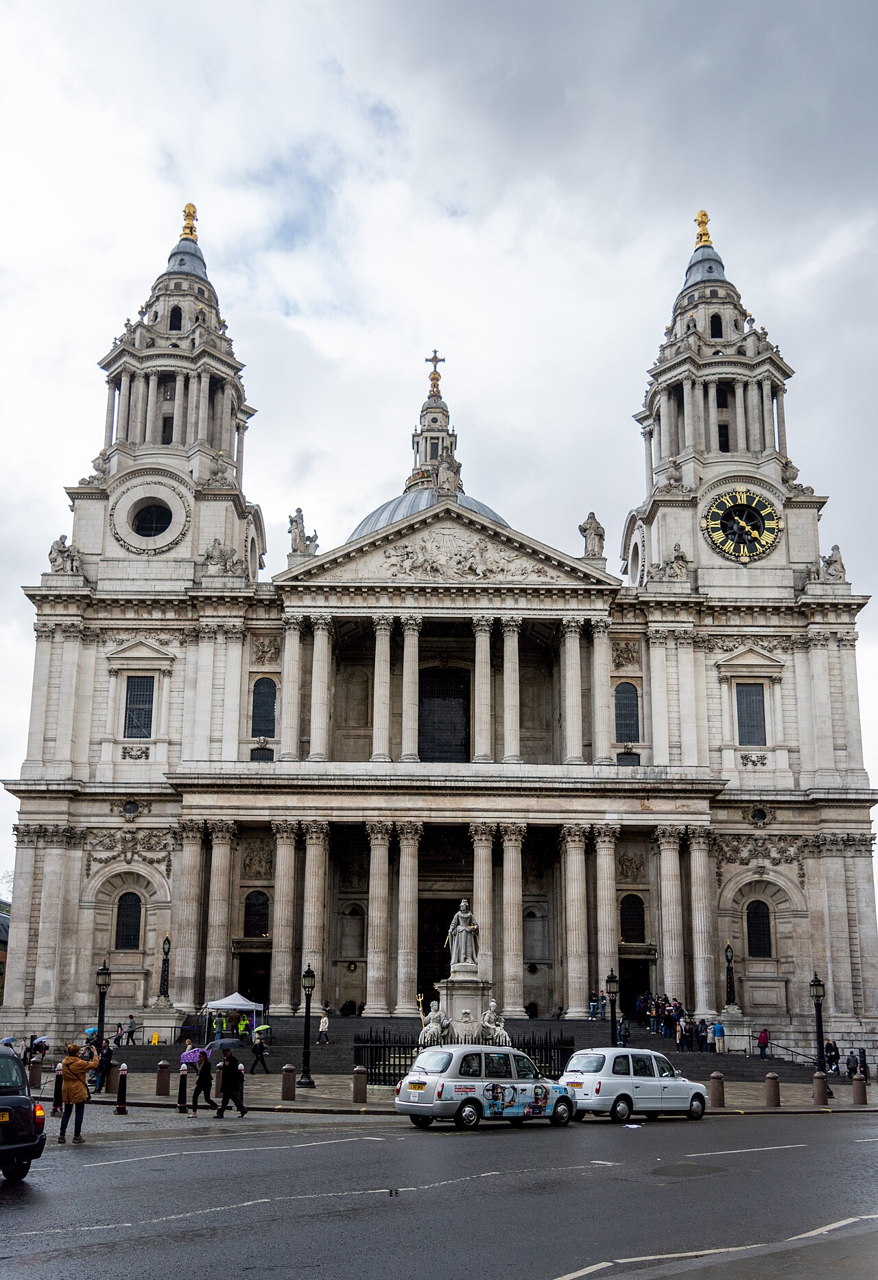 A domed baroque cathedral with two towers and a columned portico, a statue on a pedestal in front, taxis on the road.