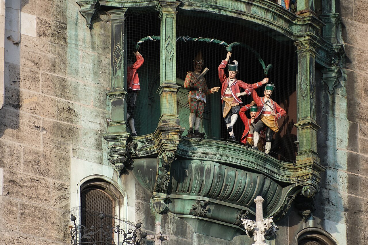 Small figures in red jackets holding green hoops in a copper bay.