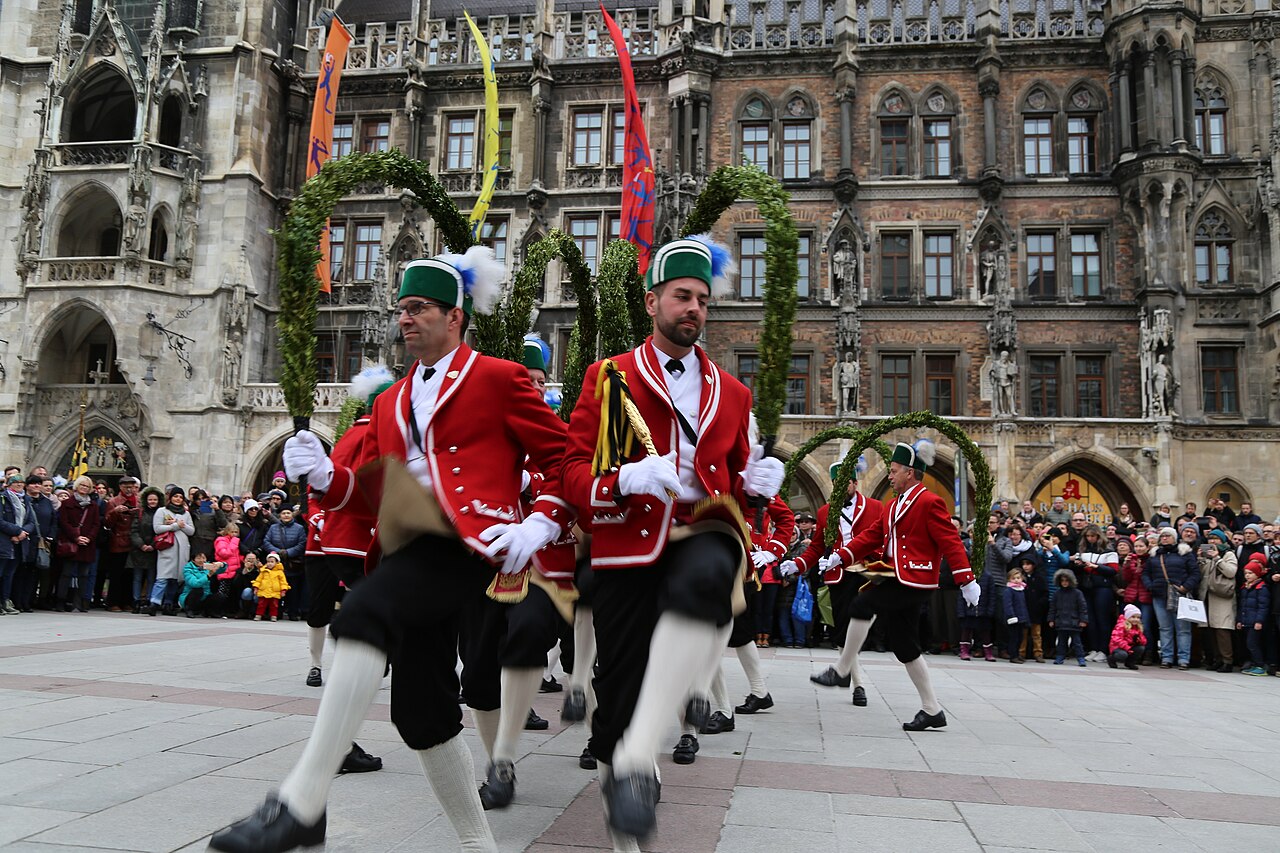 Dancers in red jackets and green caps carrying arches of green boxwood across a square.