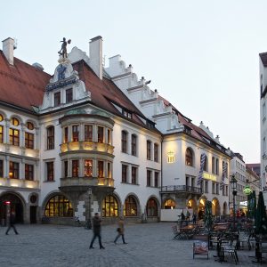 The Hofbräuhaus front on the Platzl at dusk, seen across the paved square: a white neo-Renaissance building with a stepped...