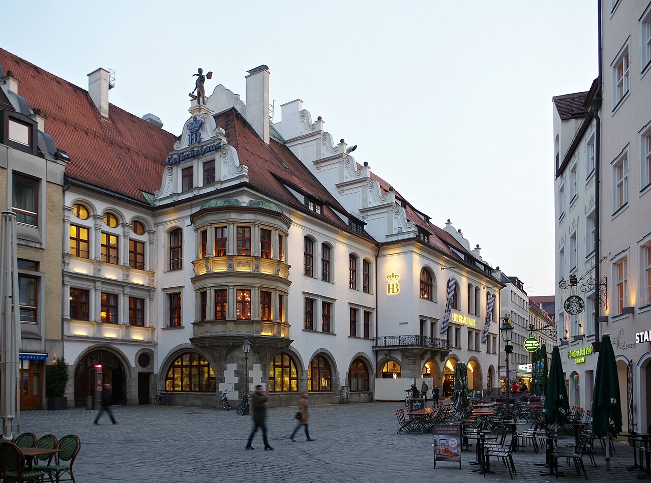 The Hofbräuhaus front on the Platzl at dusk, seen across the paved square: a white neo-Renaissance building with a stepped...