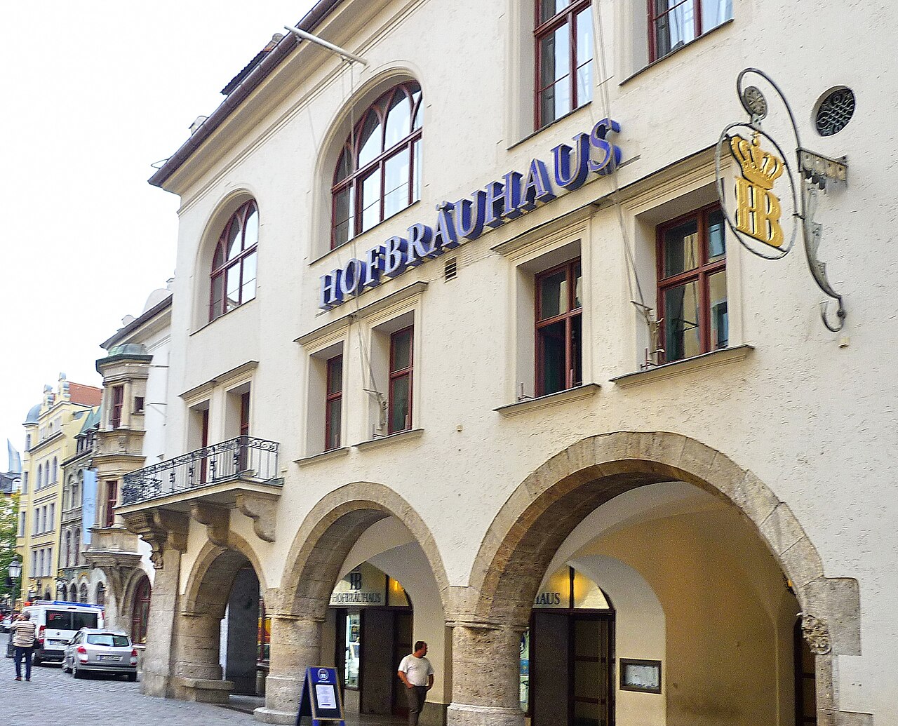 A white building with round ground-floor arches, blue lettering and a hanging sign with a crowned H B.
