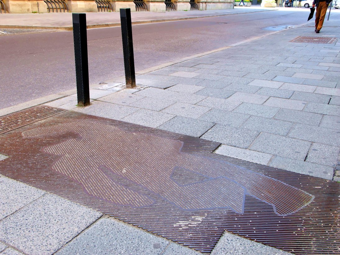 A dark ribbed metal plate set into a sidewalk between two black bollards, with the outline of a fallen figure.