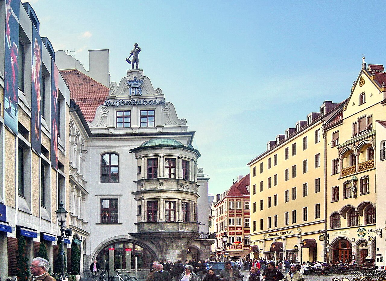 A small cobbled square with crowds of people in front of a white building with arched doorways and a corner oriel.