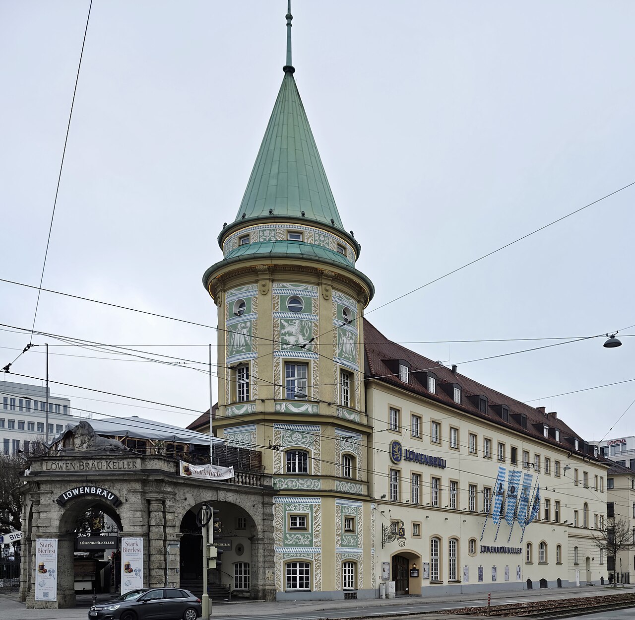 A round corner tower with a green conical roof above an arched entrance, a long hall beside it.