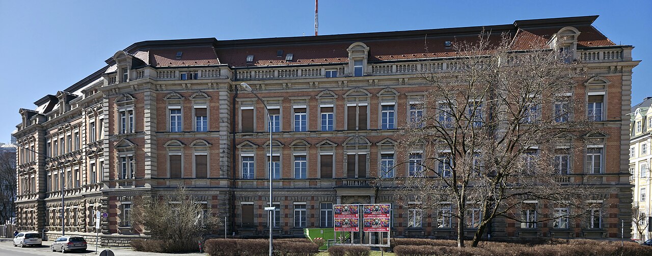A long three-story red-brick building with sandstone trim and a mansard roof behind a strip of lawn.