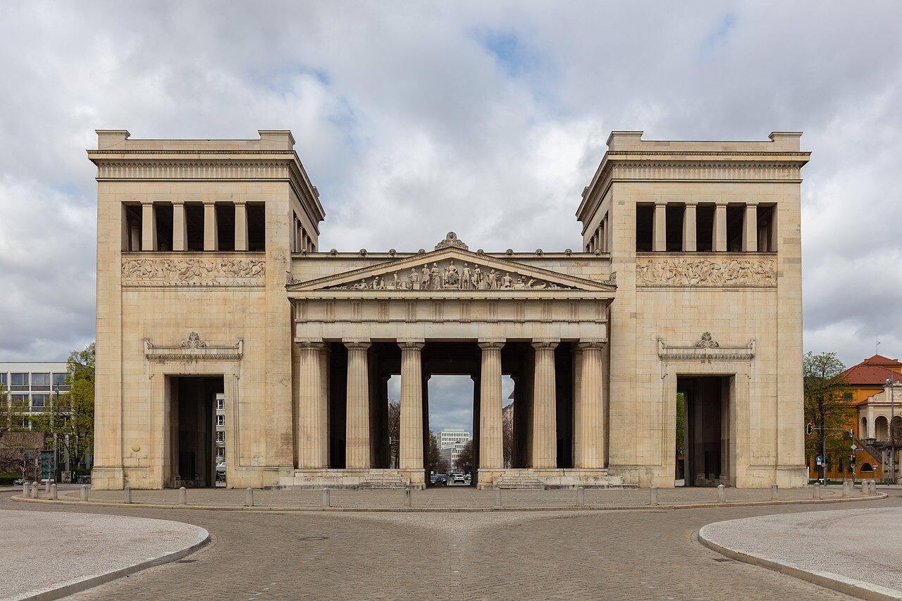 A classical gateway with columns between two tall towers, across a paved square.