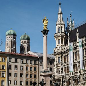 Marienplatz at street level on a sunny day: the gilded Virgin on top of the Mariensäule rises in the center, the two...