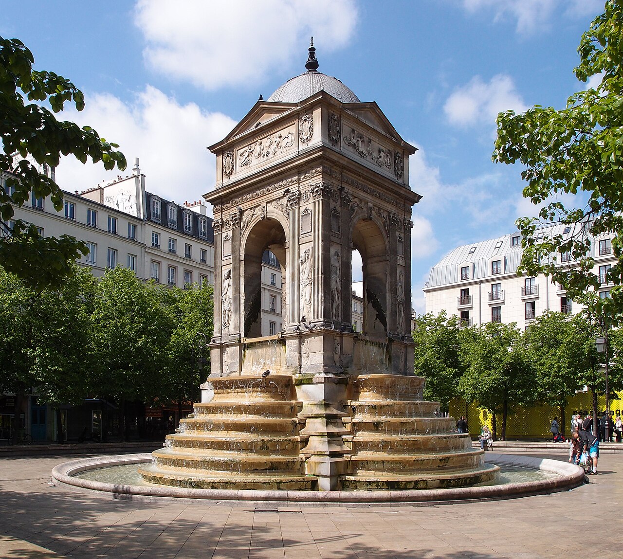 A square Renaissance fountain pavilion with arches and carved figures, on a stepped base in a paved square.
