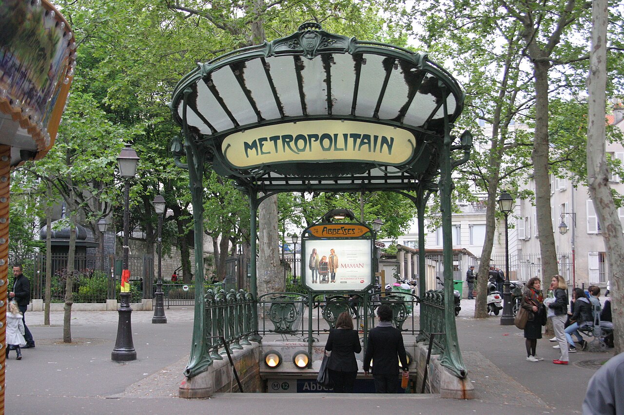 A green iron métro entrance with a fan-shaped glass roof and a yellow METROPOLITAIN sign under plane trees.