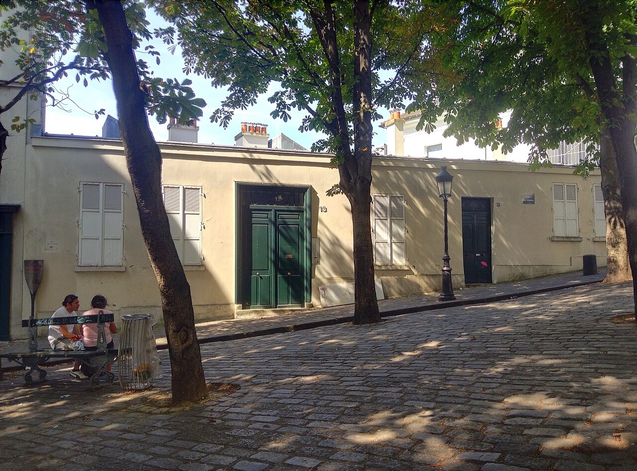 A low pale building with shuttered windows and a green double door on a cobbled square under trees.
