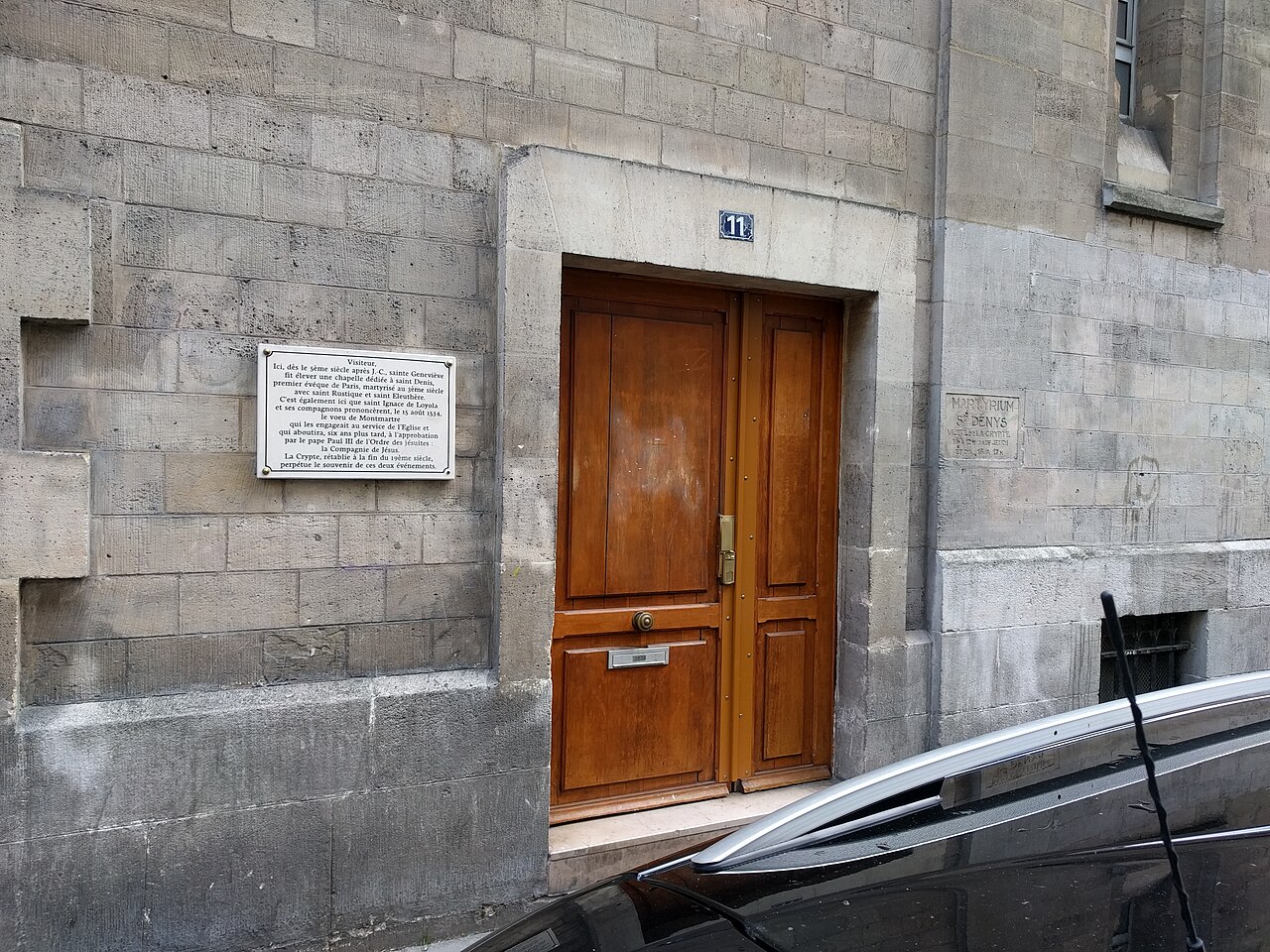 A stone wall with a wooden double door numbered 11, a white plaque on the left and carved letters on the right.
