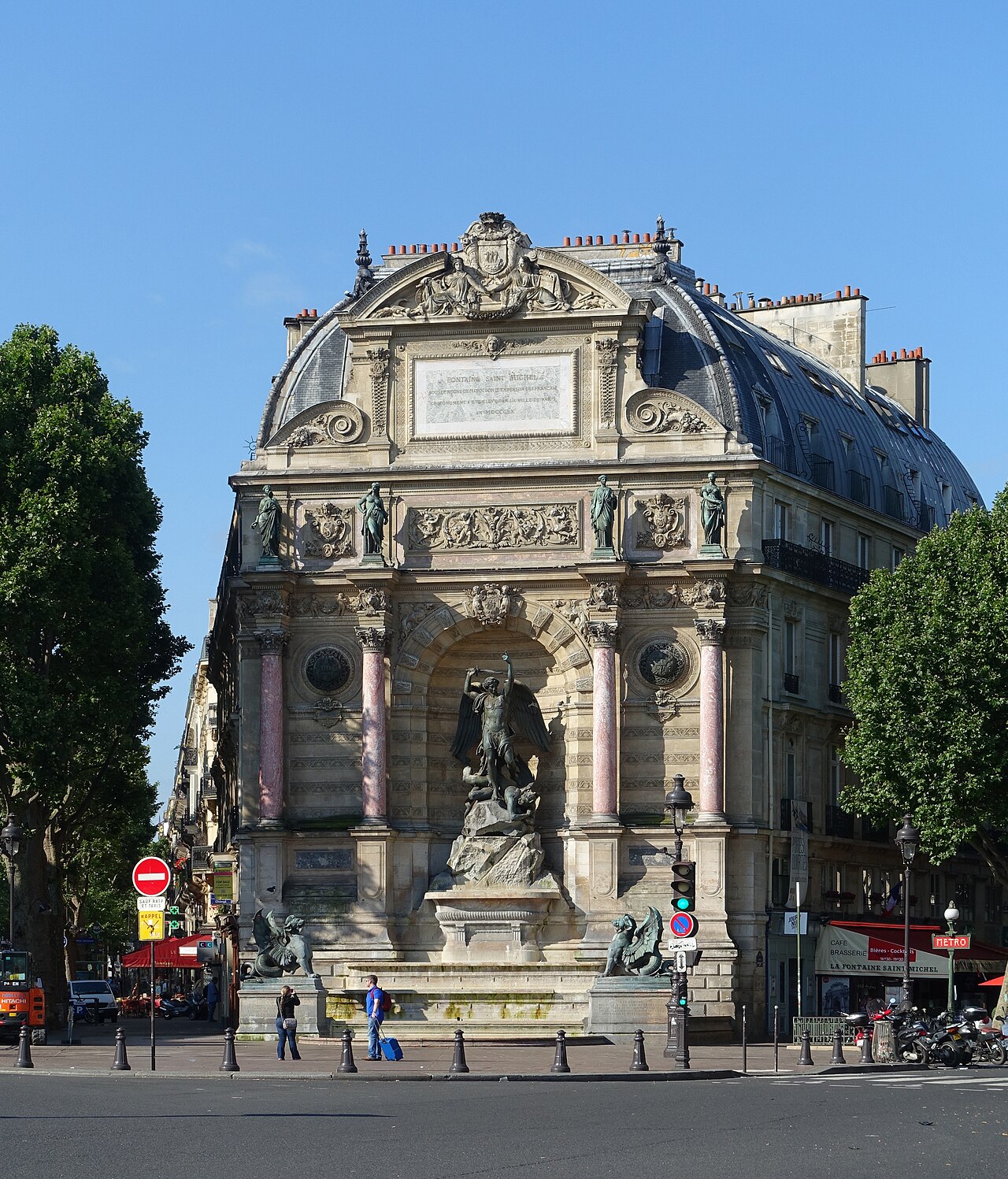 A tall fountain in a triumphal arch on a corner building, with a winged saint over a devil and two dark winged beasts before the basin.
