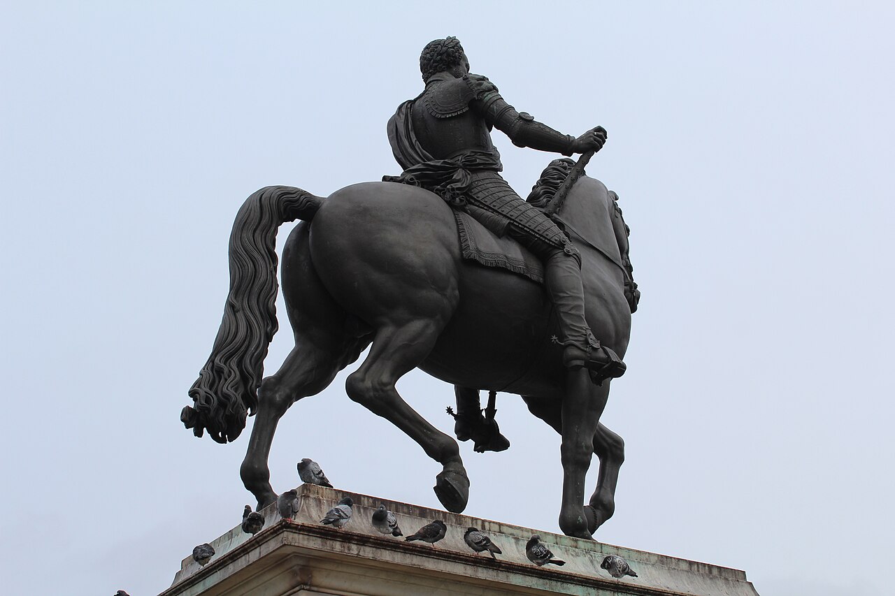 A dark bronze king on a prancing horse on a stone pedestal, against a pale sky.
