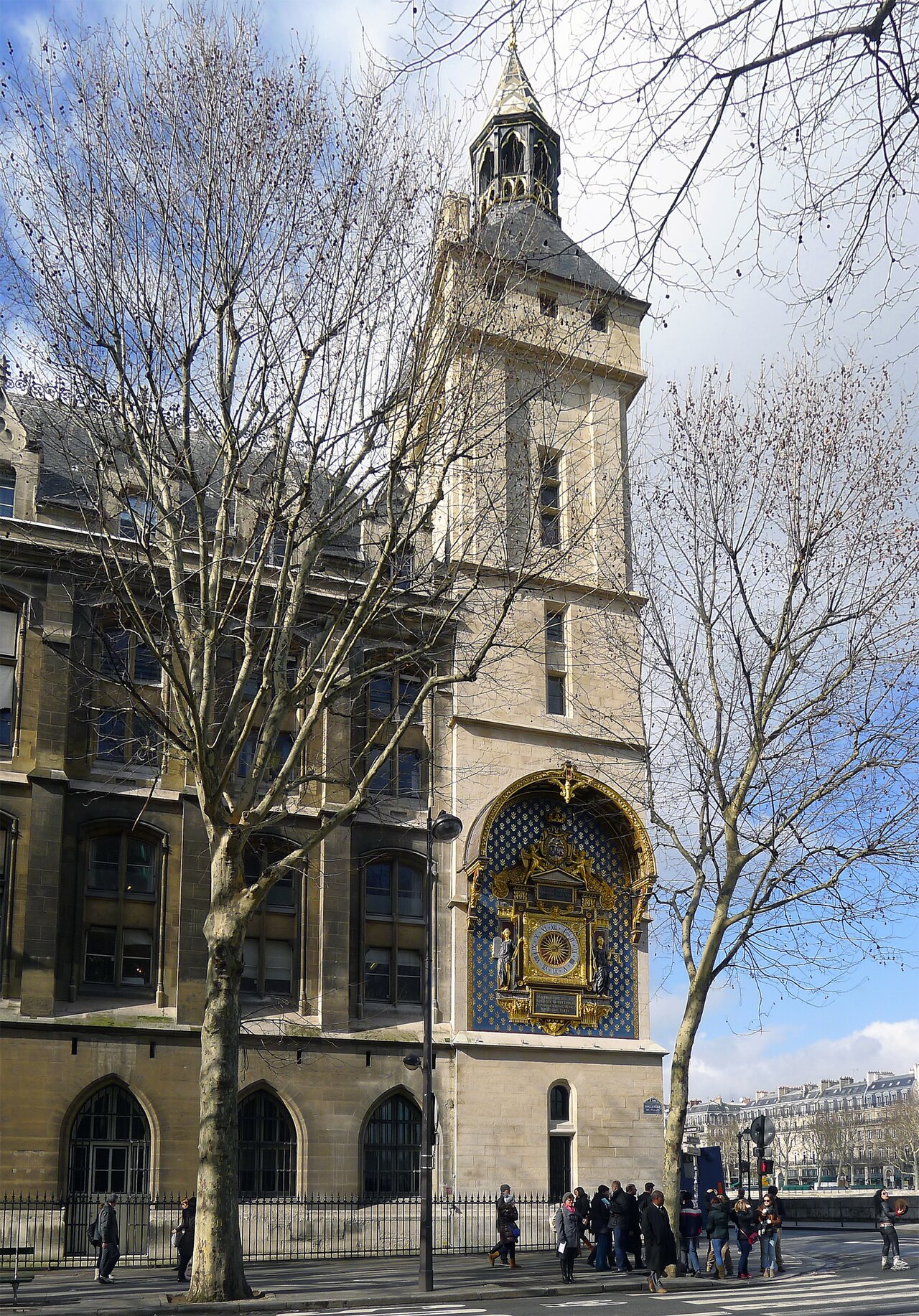 A tall square stone tower with a blue and gold clock face on its corner, behind bare trees.