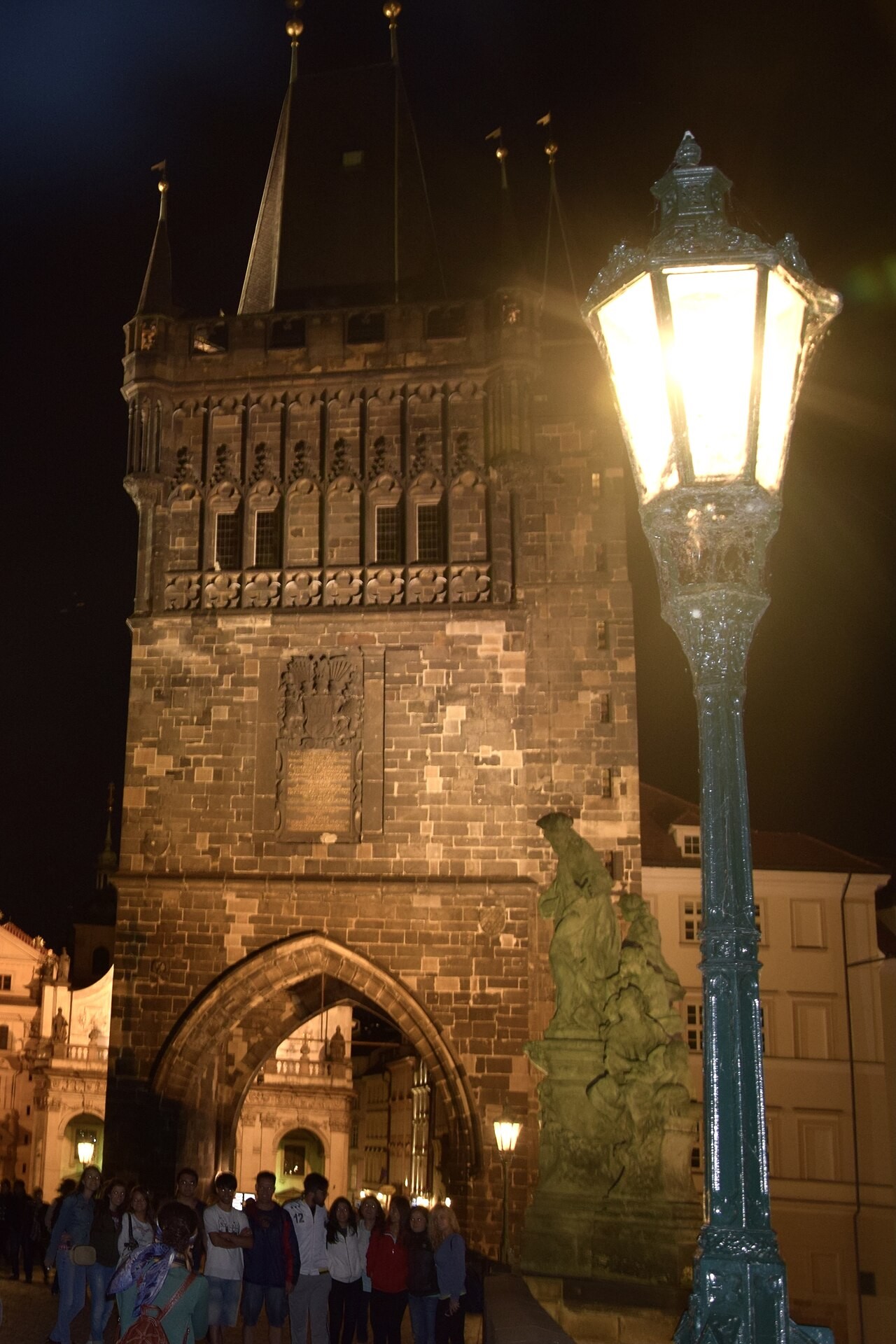 The floodlit gate tower behind a lit street lamp, with people under the arch.