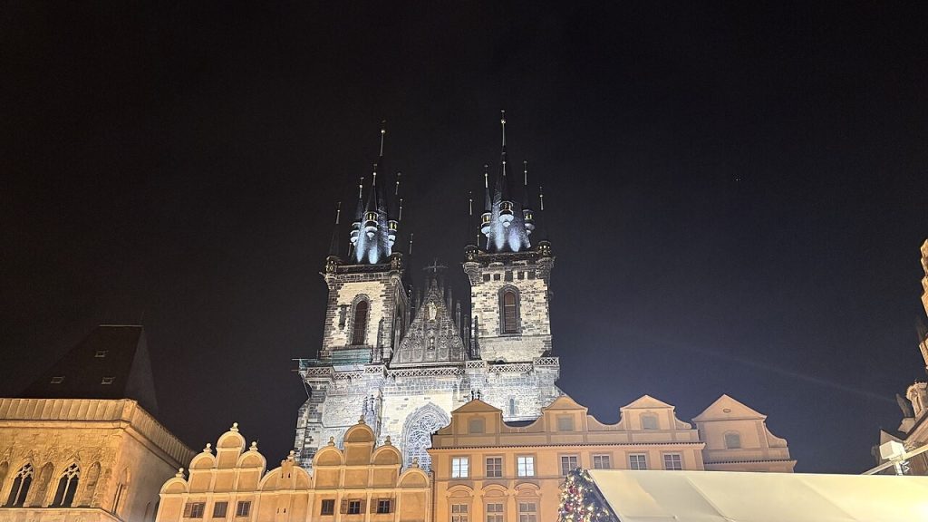 Old Town Square at night: the floodlit twin spires of Týn Church rising behind the lit gables of the houses on the square...