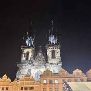 Old Town Square at night: the floodlit twin spires of Týn Church rising behind the lit gables of the houses on the square...