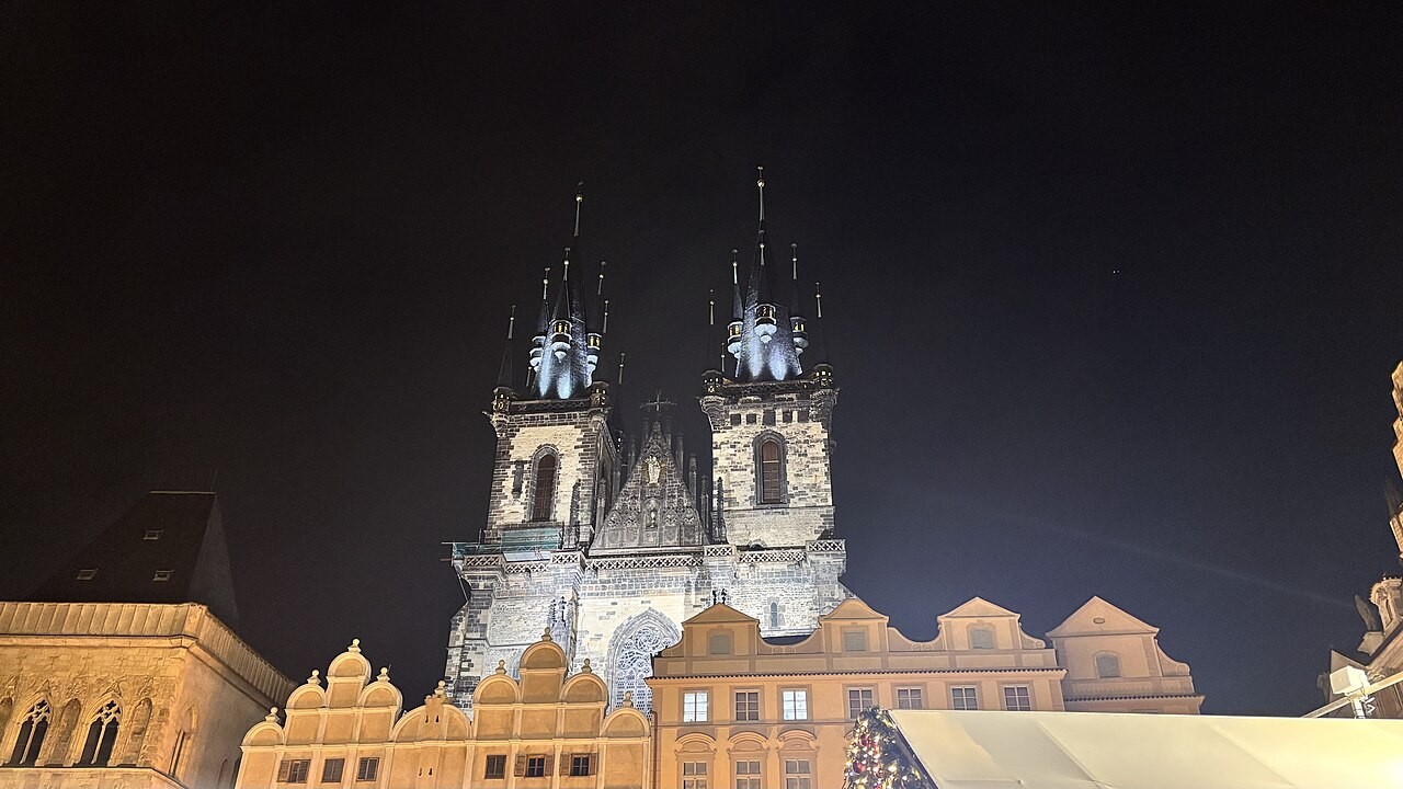 Old Town Square at night: the floodlit twin spires of Týn Church rising behind the lit gables of the houses on the square...
