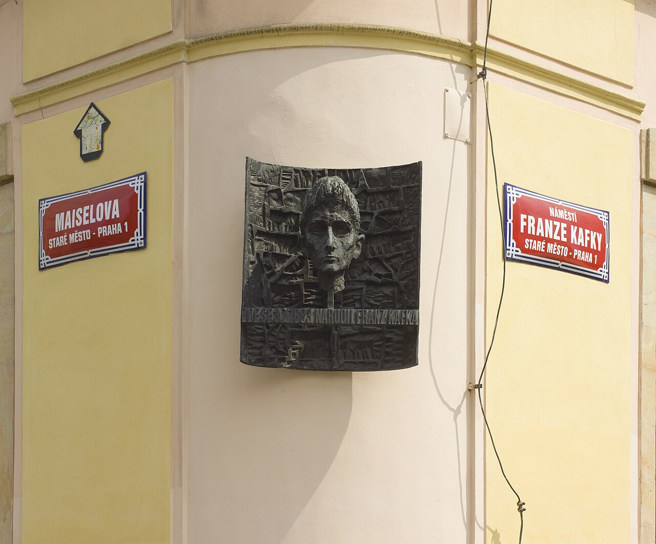 A rounded yellow building corner with a dark bronze relief of a man's head between two red street signs.