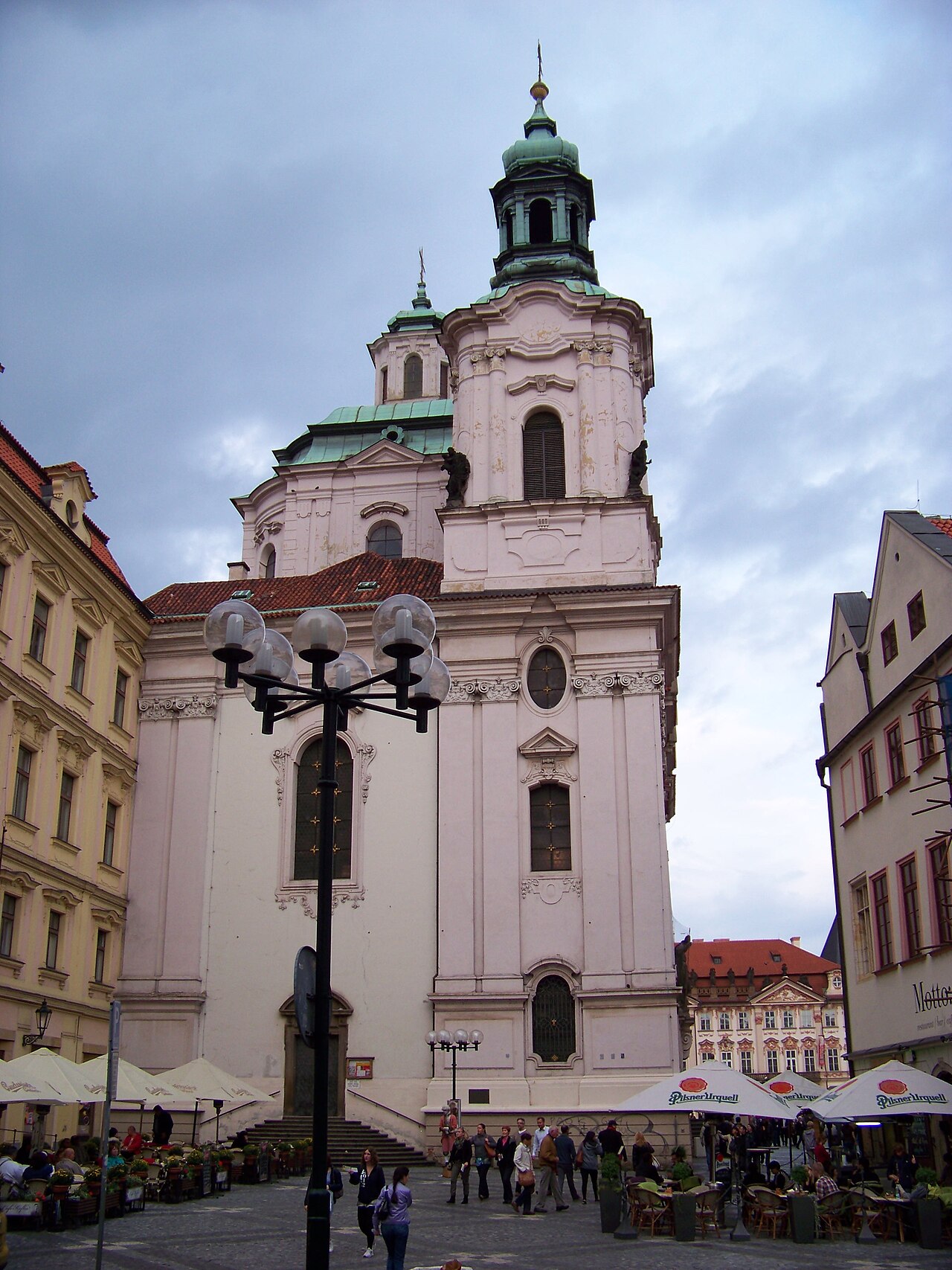 A white Baroque church with a green-domed tower above café umbrellas.