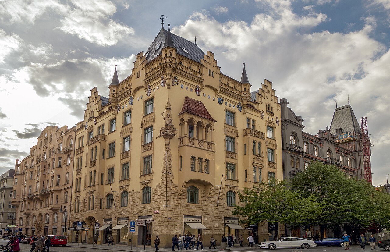 An ochre corner apartment block with turrets, gables and an oriel above shop awnings.