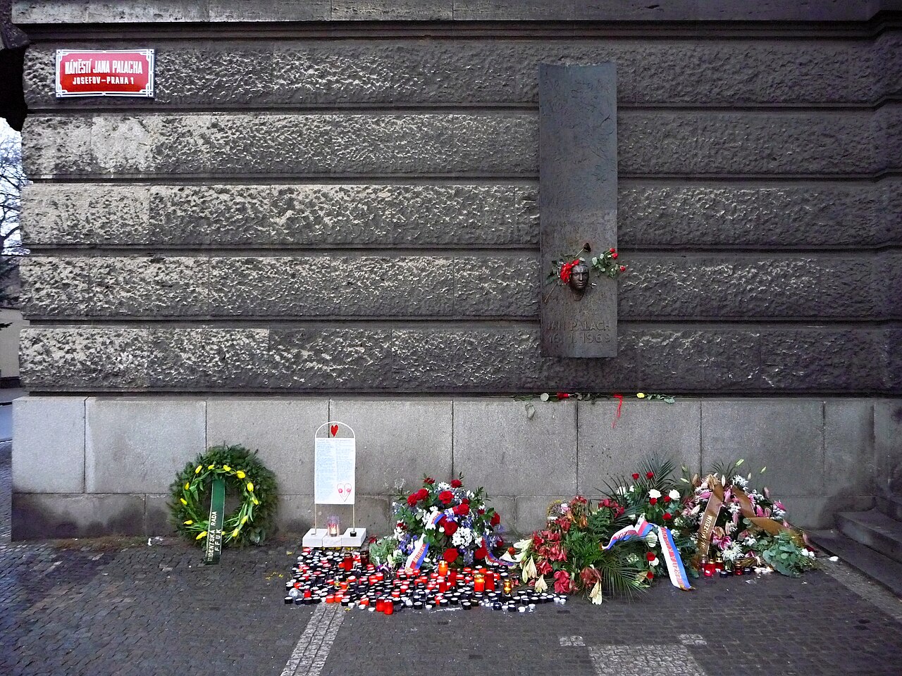 A stone building corner with a street sign and a memorial set into the wall, flowers below.