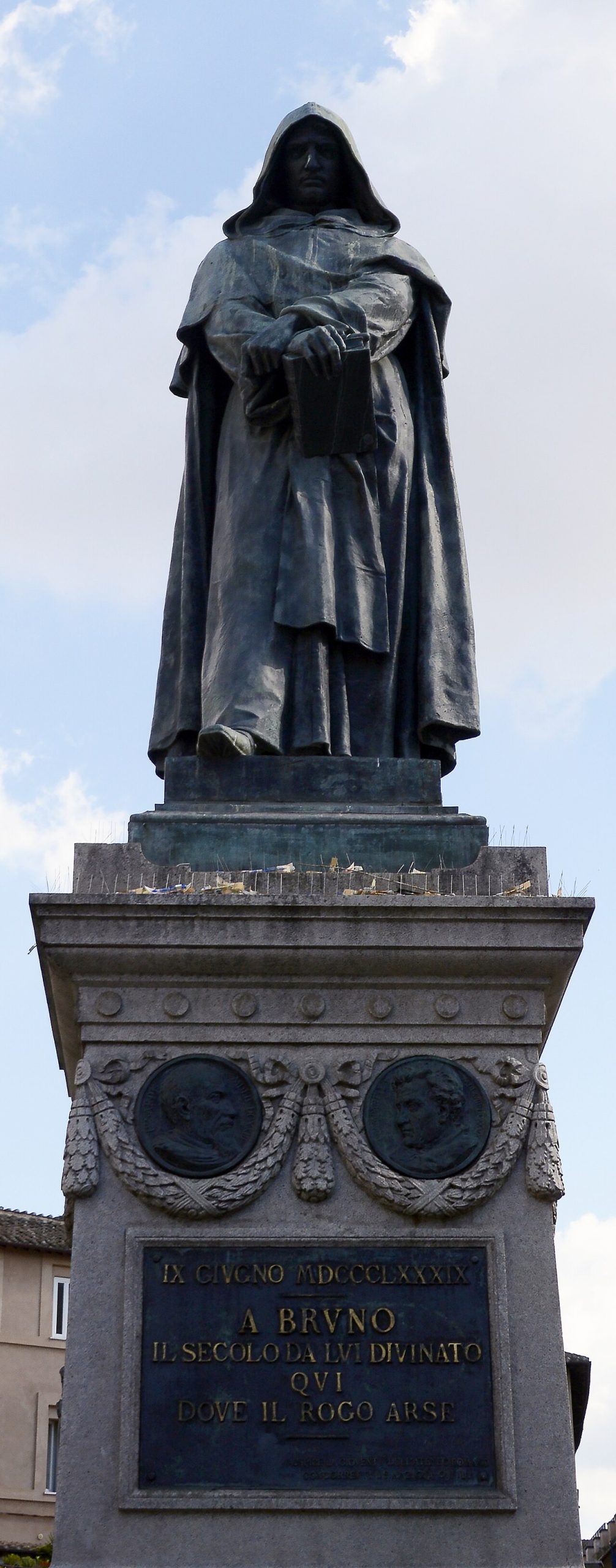 A hooded bronze figure holding a book, on a tall stone pedestal, seen from below.