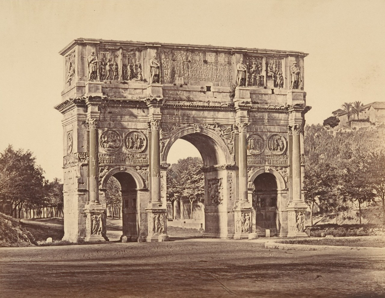 A sepia photograph of a triple stone arch with columns, round reliefs and standing statues on top, trees behind.