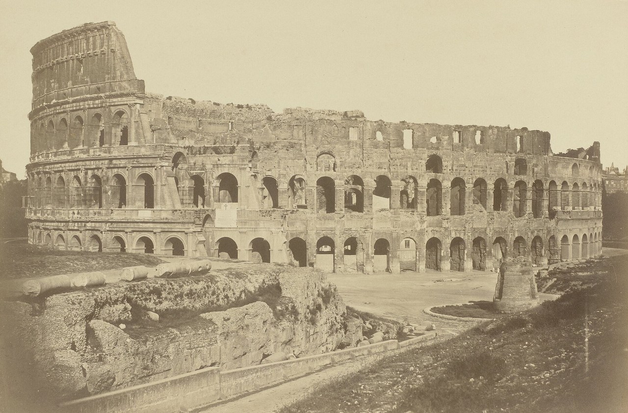 A sepia photograph of the Colosseum's tall outer wall ending at a buttress on the left, the lower inner ring beyond, and a cone-shaped ruin in front on the right.