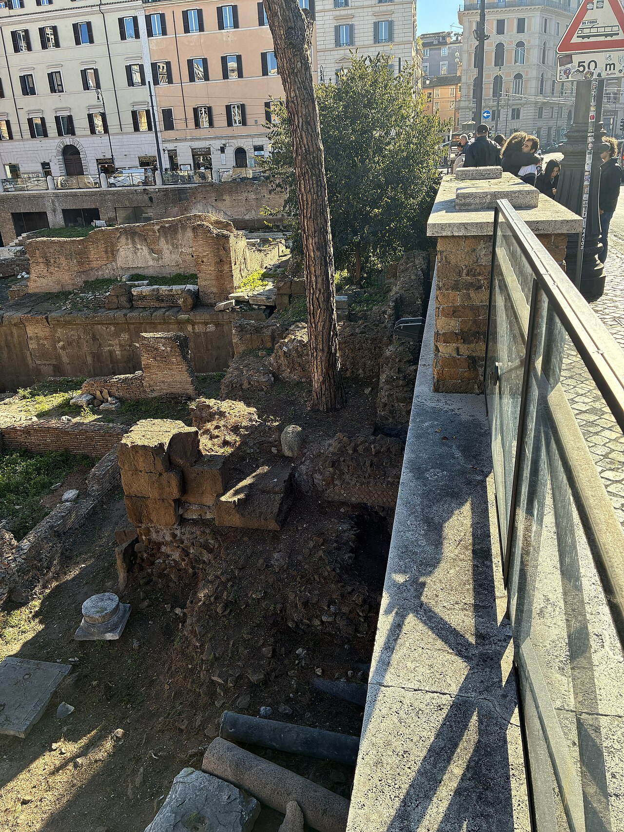 Tuff blocks and brick walls with fallen column drums and a pine trunk below a glass and stone parapet.