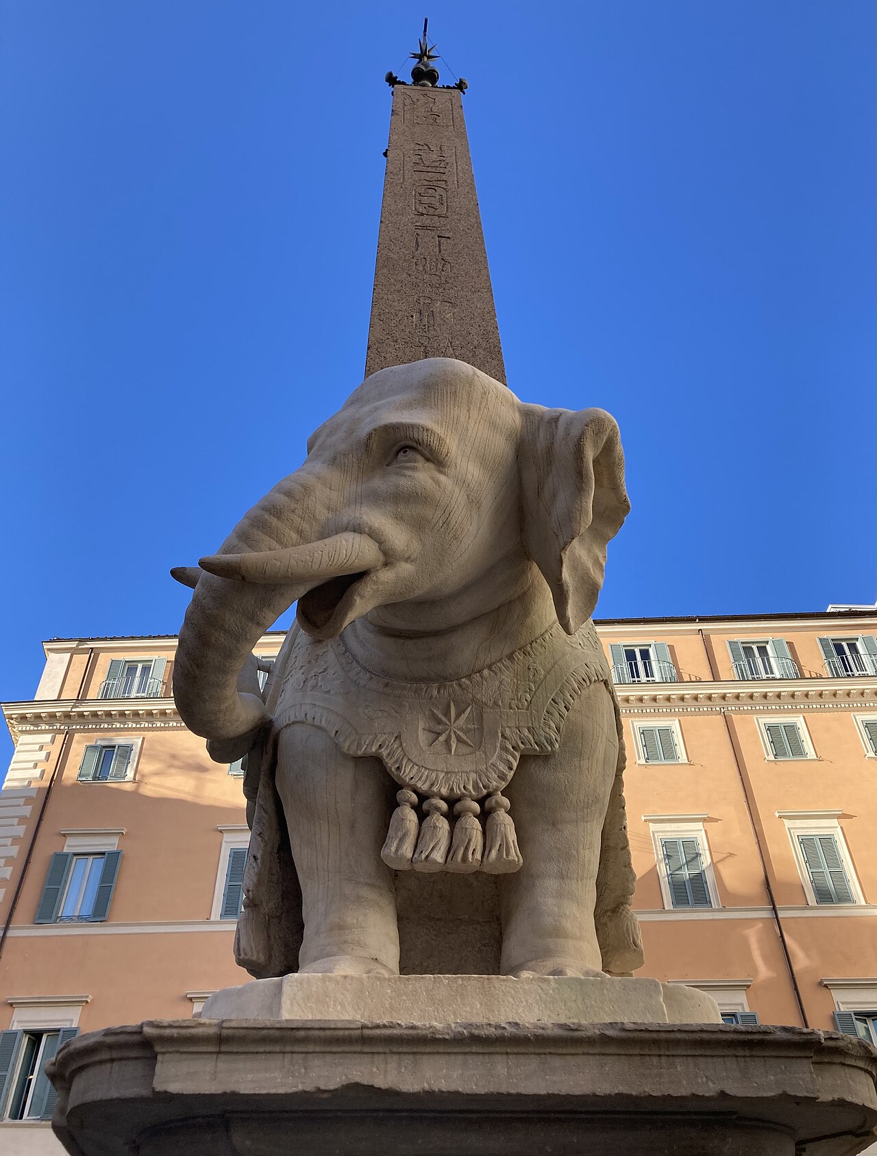 A small marble elephant wearing a fringed saddle cloth, carrying a slim obelisk, against a blue sky.