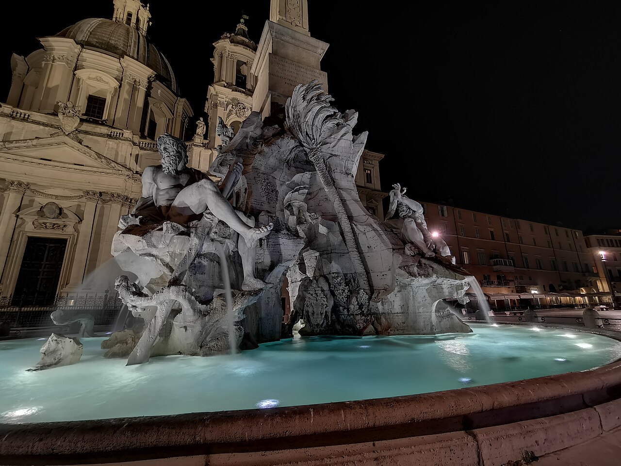A large baroque fountain with marble river gods on rocks and a lit basin, a domed church behind.