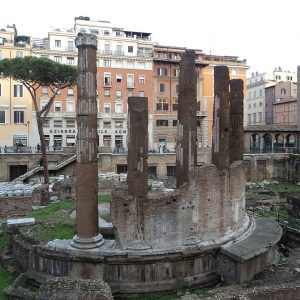 A sunken field of ruins with a round temple base and tall standing columns, apartment houses and a medieval tower beyond.