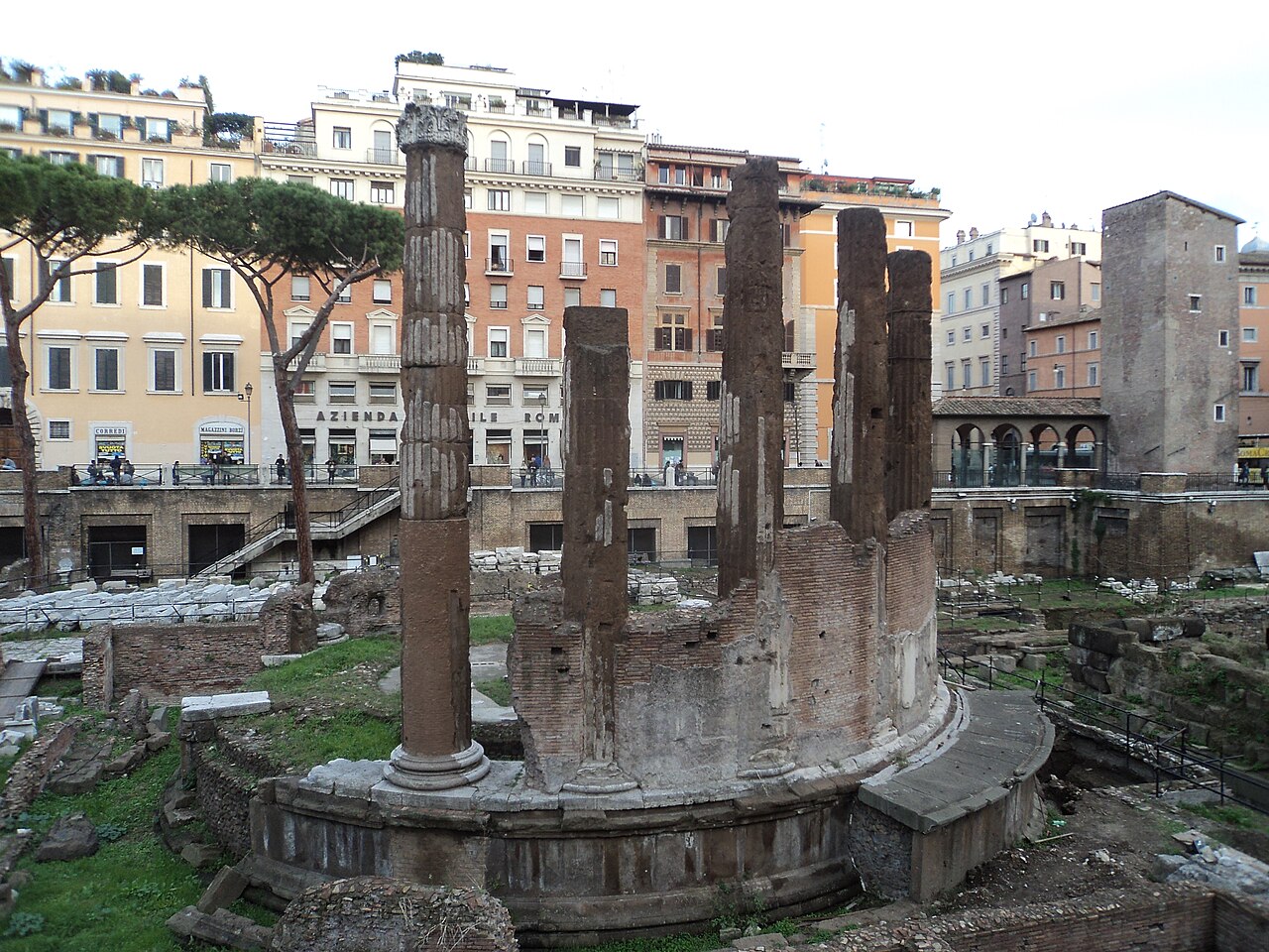 A sunken field of ruins with a round temple base and tall standing columns, apartment houses and a medieval tower beyond.