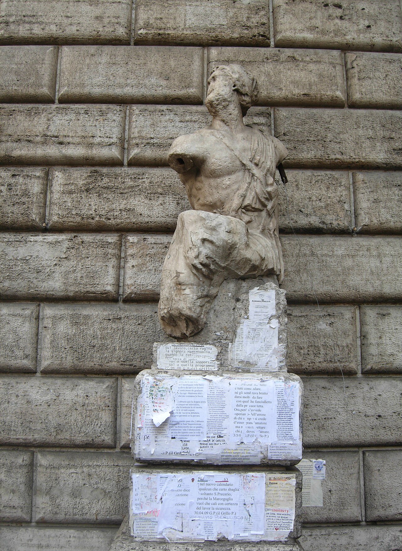 A worn marble torso with a battered head on a stone pedestal covered with pinned papers.