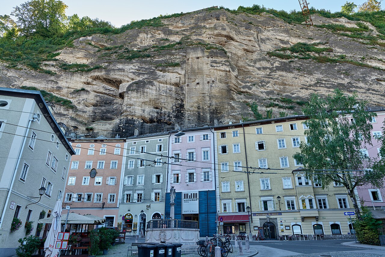 A row of narrow pastel house fronts at the foot of the Mönchsberg, the bare overhanging conglomerate rock face rising directly above their roofs and f