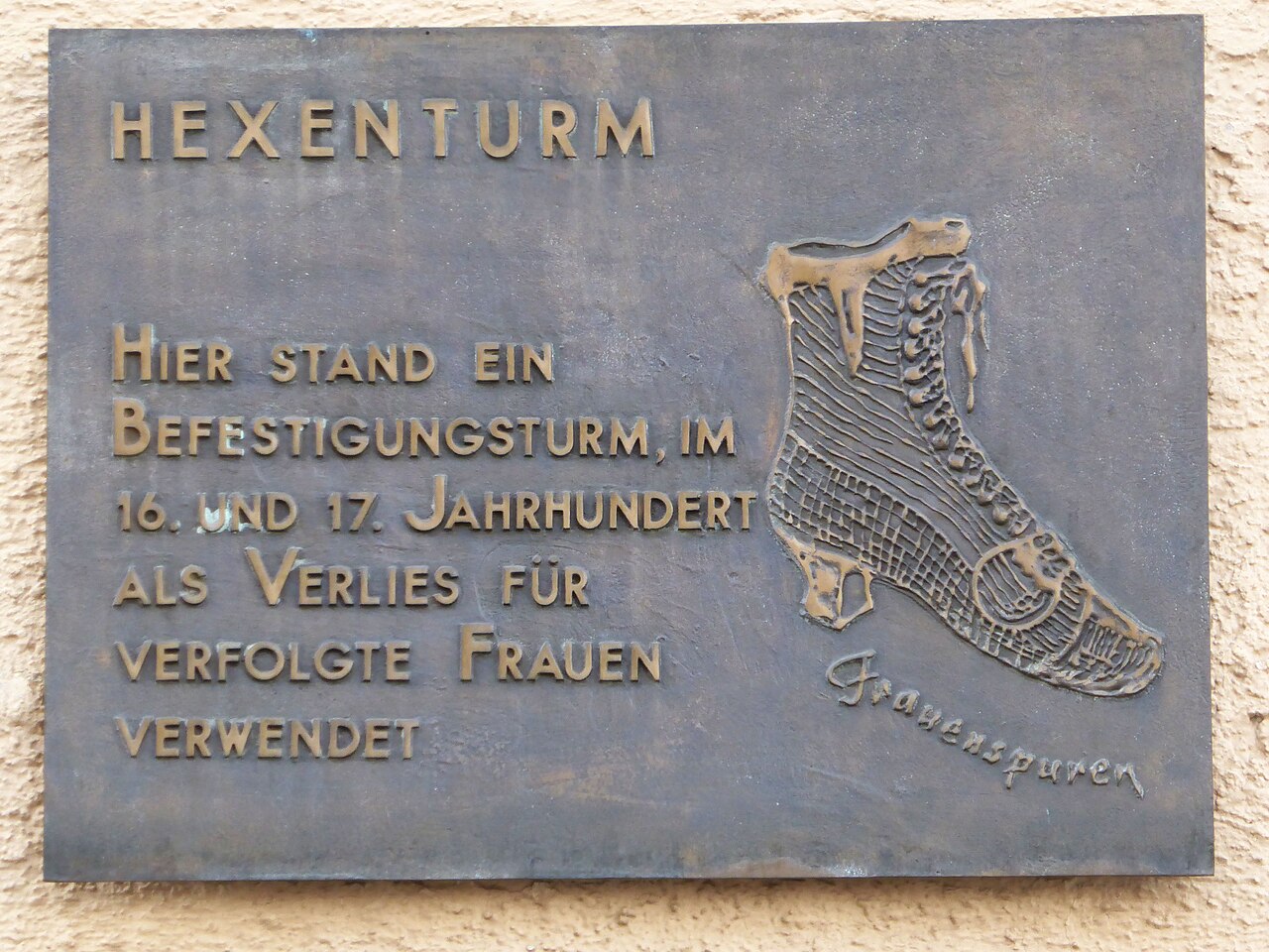 A dark bronze plaque with raised letters: 'HEXENTURM / HIER STAND EIN BEFESTIGUNGSTURM, IM 16.