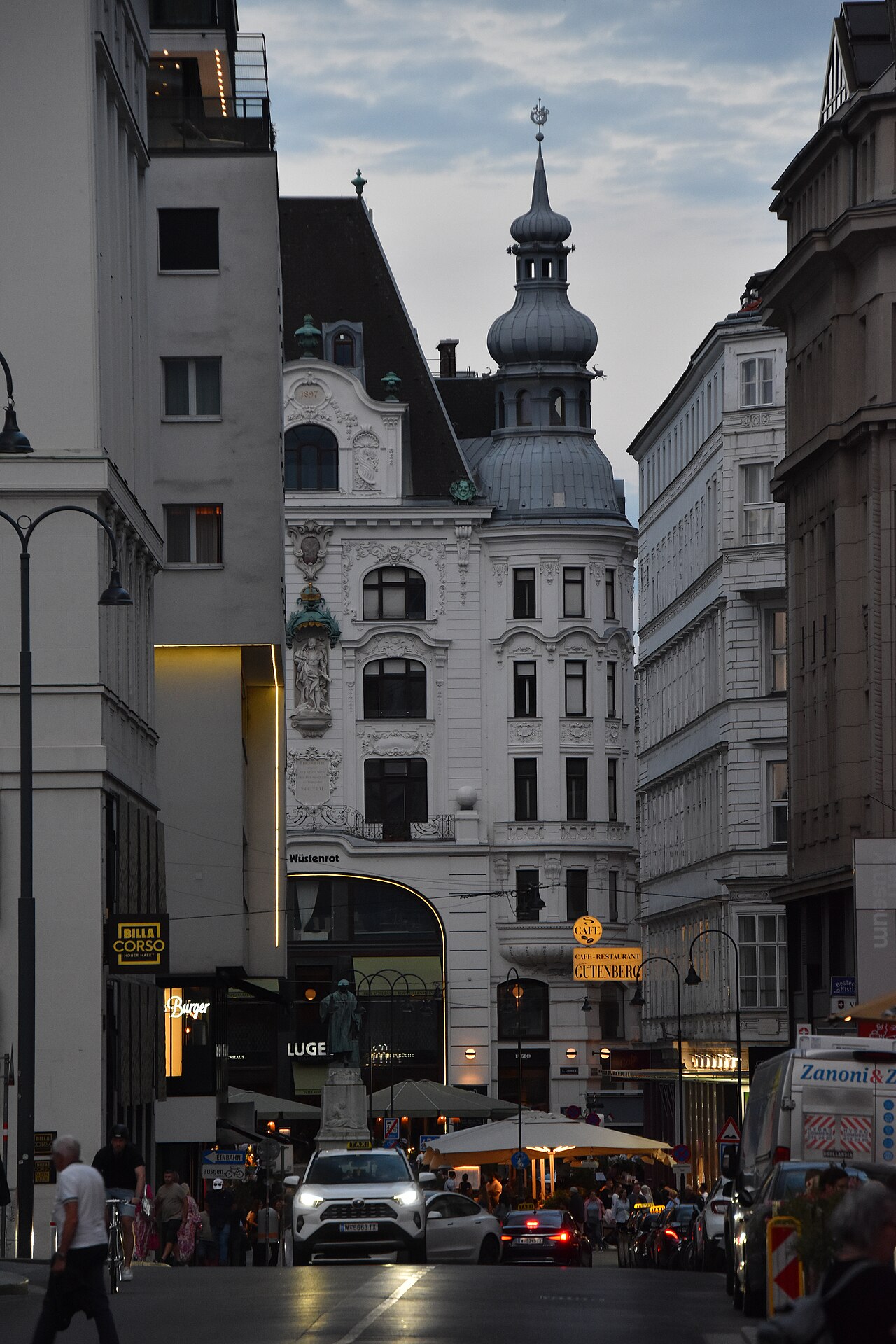 A city square at dusk with tall buildings, lit windows and street lamps.