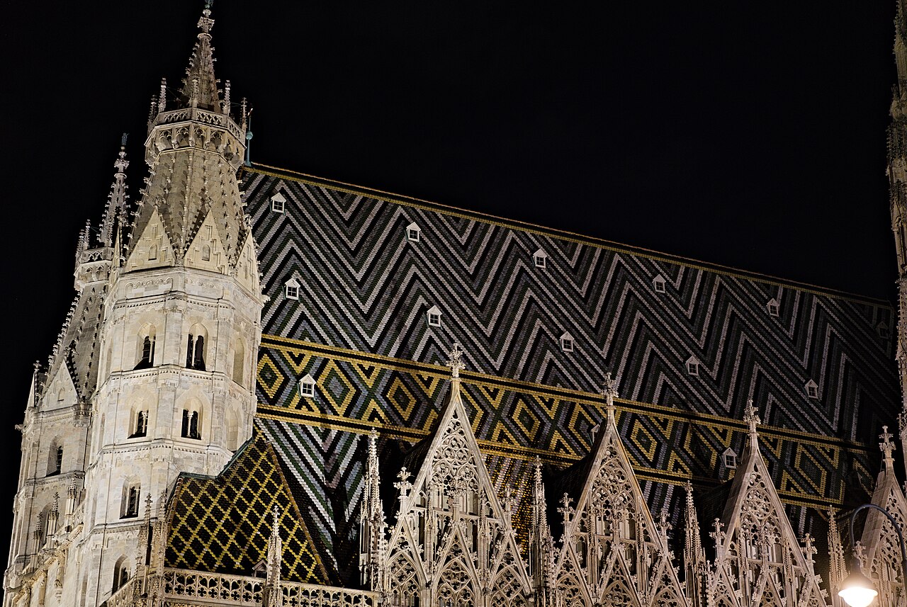 The long Gothic wall of a cathedral lit gold against a black sky, with a steep patterned roof above.
