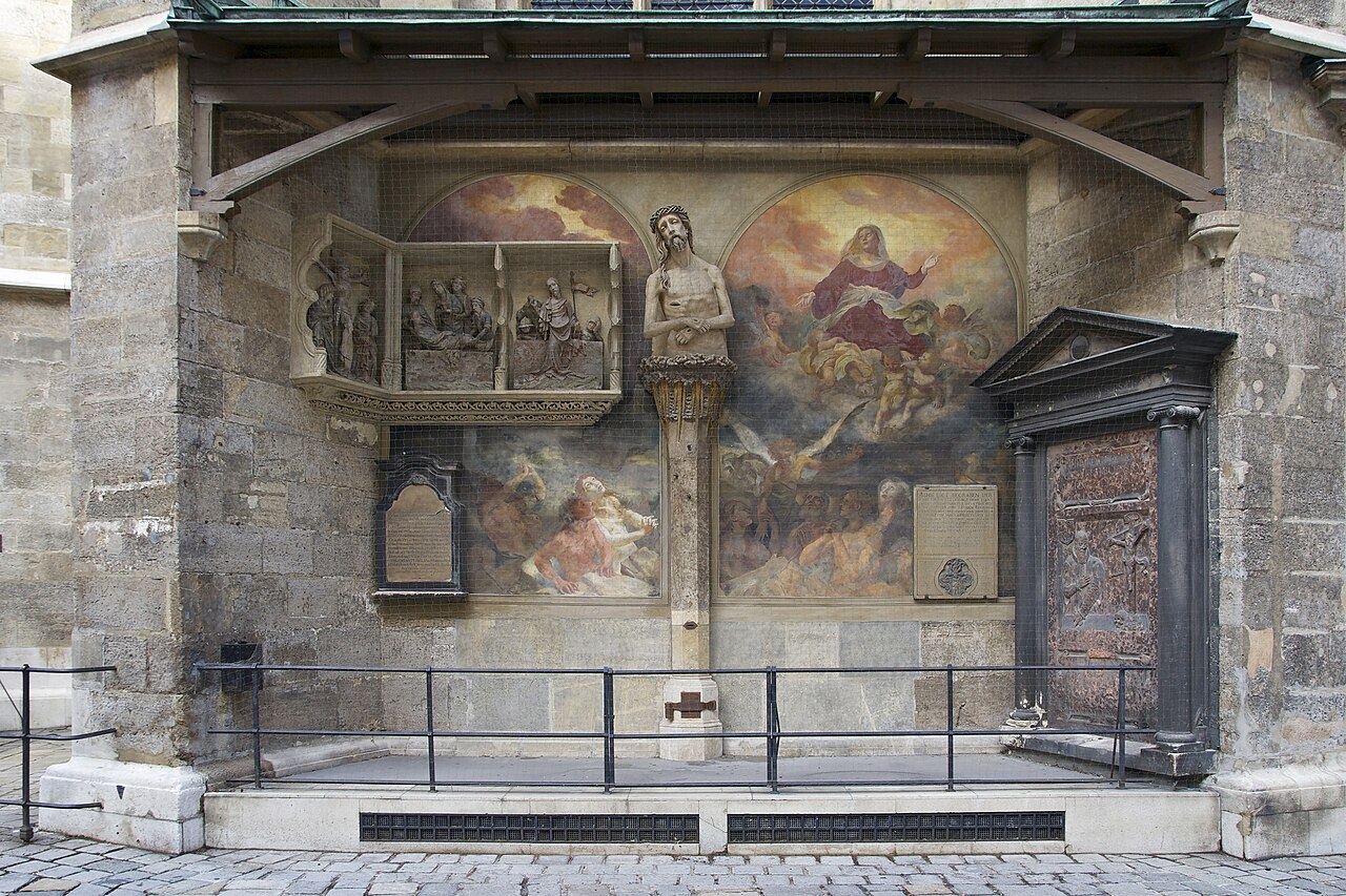 A stone half figure of Christ with a crown of thorns in a small roofed niche behind an iron railing.